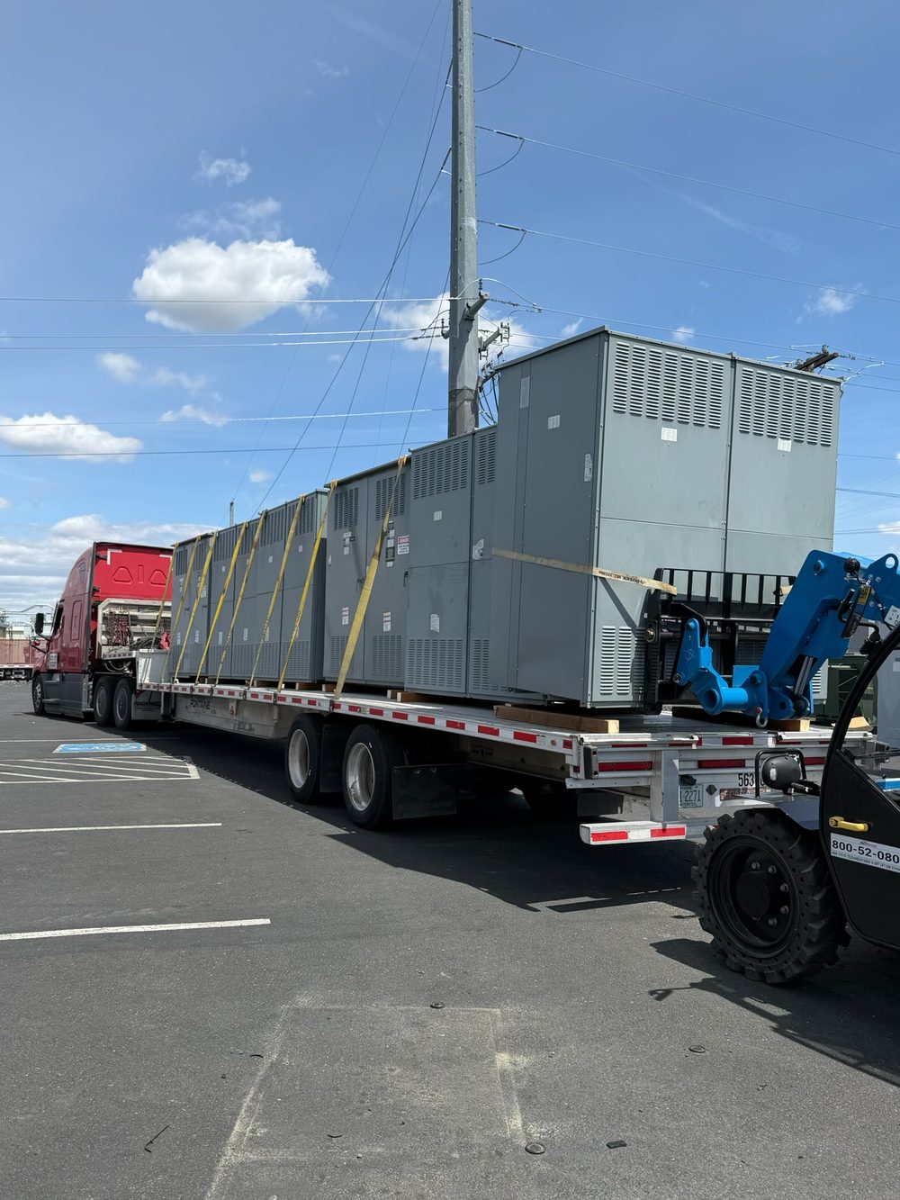 Multiple used switchgear sections strapped down across a flatbed trailer with a telehandler standing by, loaded out for nationwide trucking